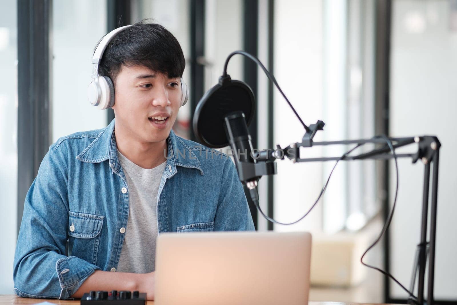 A man wearing headphones and a blue shirt is sitting in front of a laptop. He is wearing a microphone and he is recording something