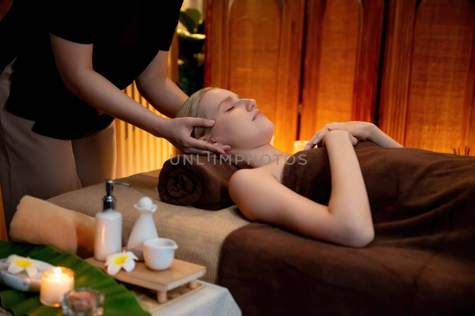 Caucasian woman enjoying relaxing anti-stress head massage and pampering facial beauty skin recreation leisure in warm candle lighting ambient salon spa in luxury resort or hotel. Quiescent