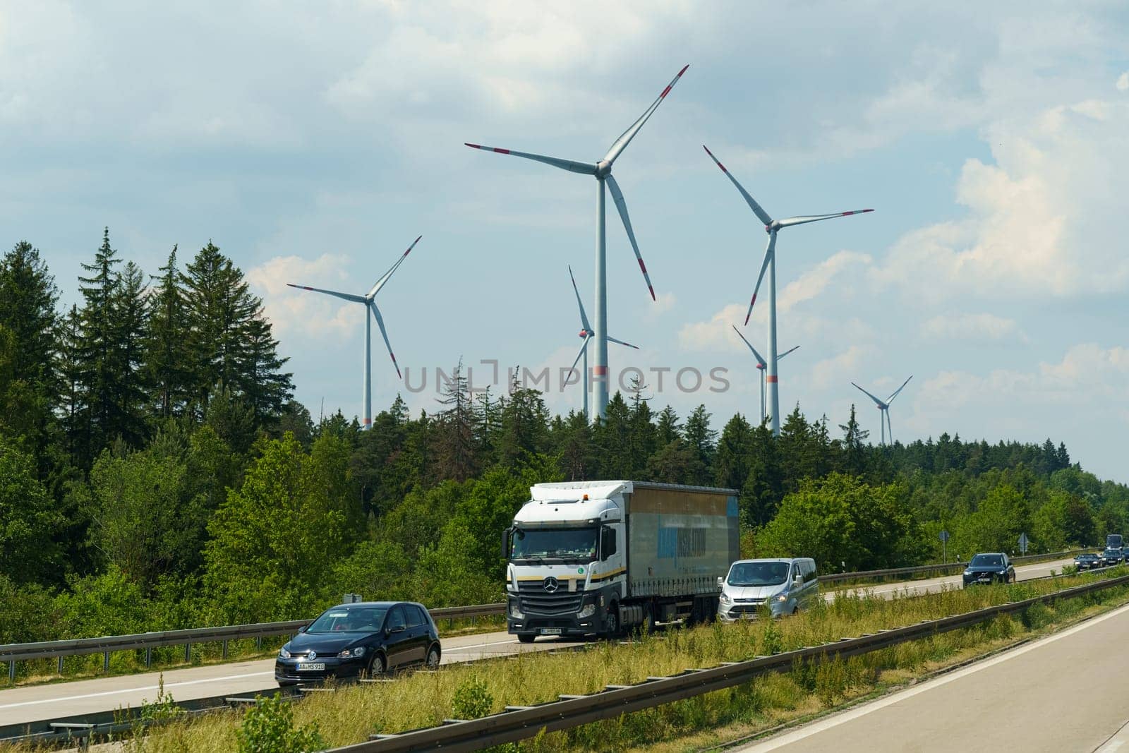Kassel, Germany - June 9, 2023: A truck drives down a road flanked by tall wind turbines. The massive blades of the turbines spin in the wind as the truck moves along the highway.