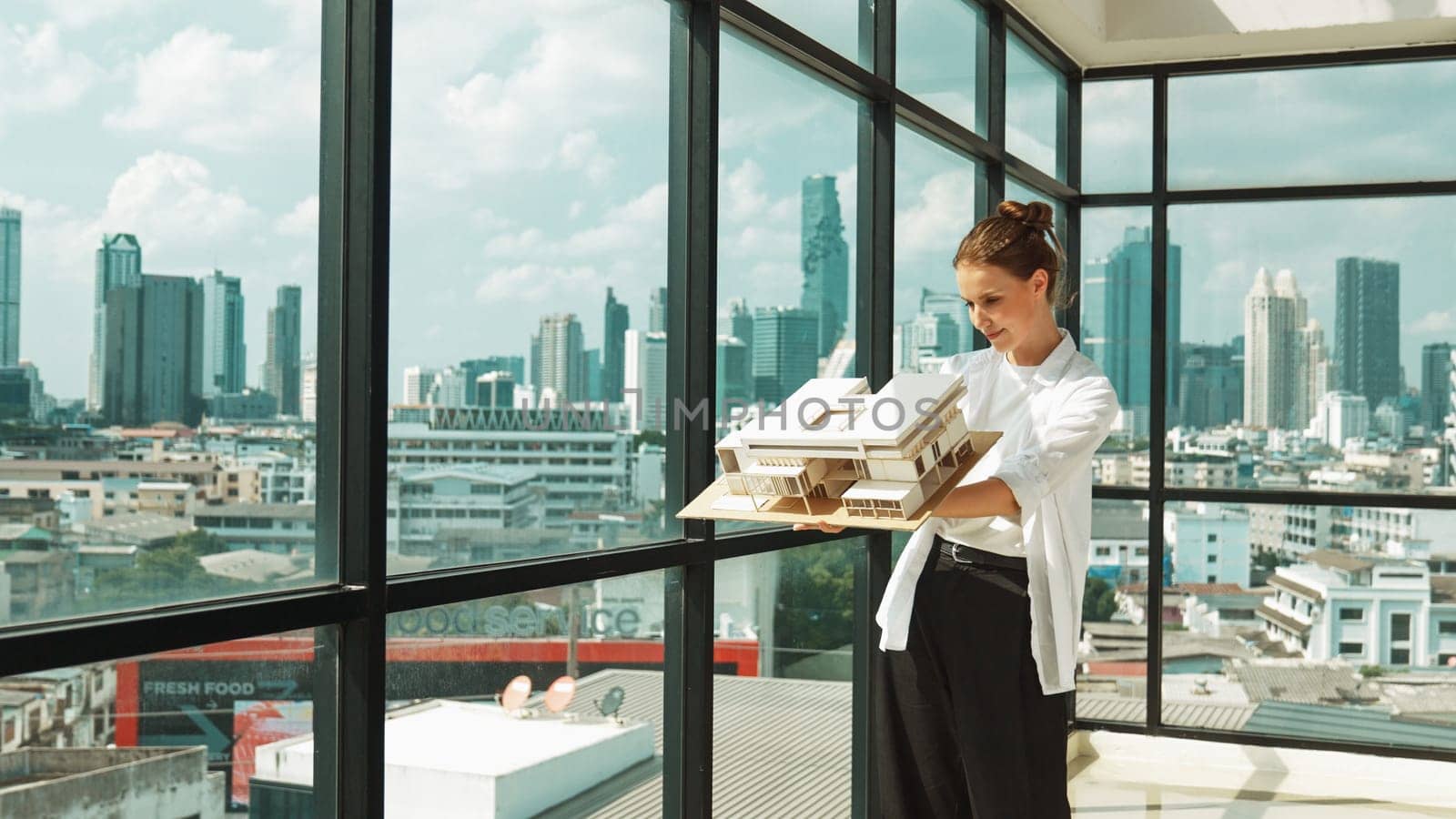 Young smart caucasian beautiful architect engineer raised house model while inspect carefully surrounded by city skyscraper view. Civil engineering, architectural studio. project planing. Tracery