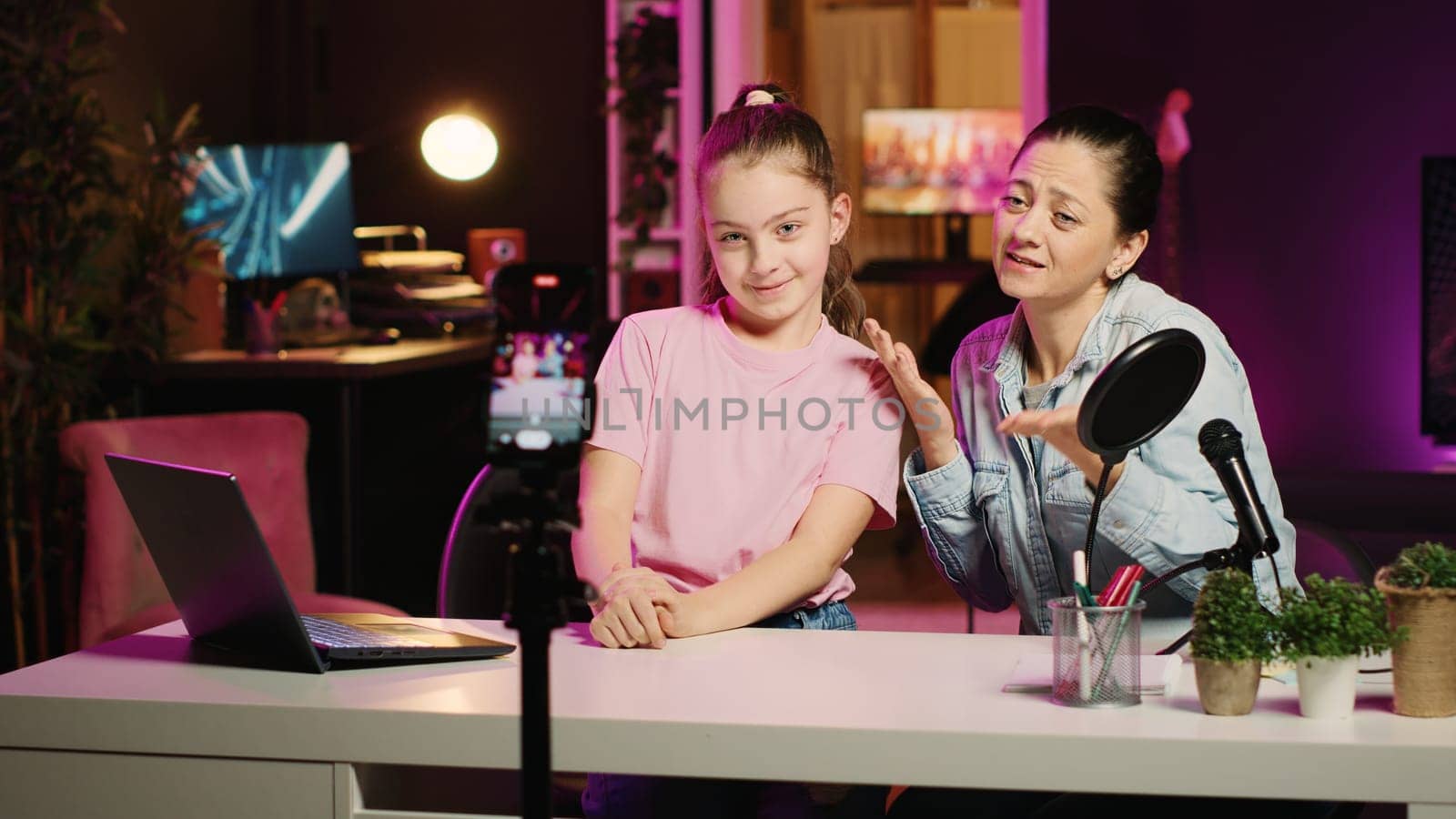Young girl and her mother filming content for their internet channel, talking about their relationship. Cute kid and mom recording family vlog in apartment studio with pink neon lighting as background