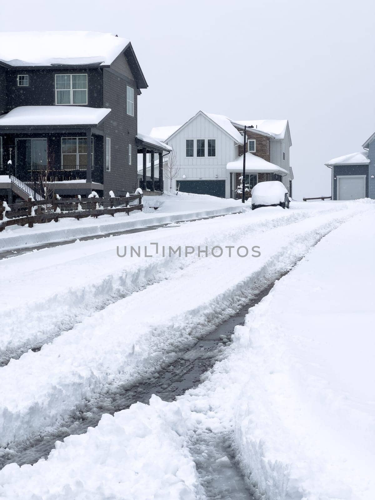 The snow-covered street in a residential area features a driveway and sidewalk cleared of snow, leading to a cozy house. The muted colors of the homes stand out against the blanket of white snow.