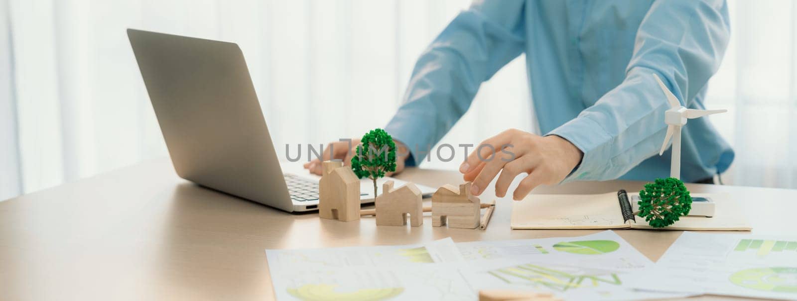 Professional architect plans to use green design in an eco house. Skilled engineer design an eco house blueprint by using laptop on table with house and windmill model scatter around. Delineation.