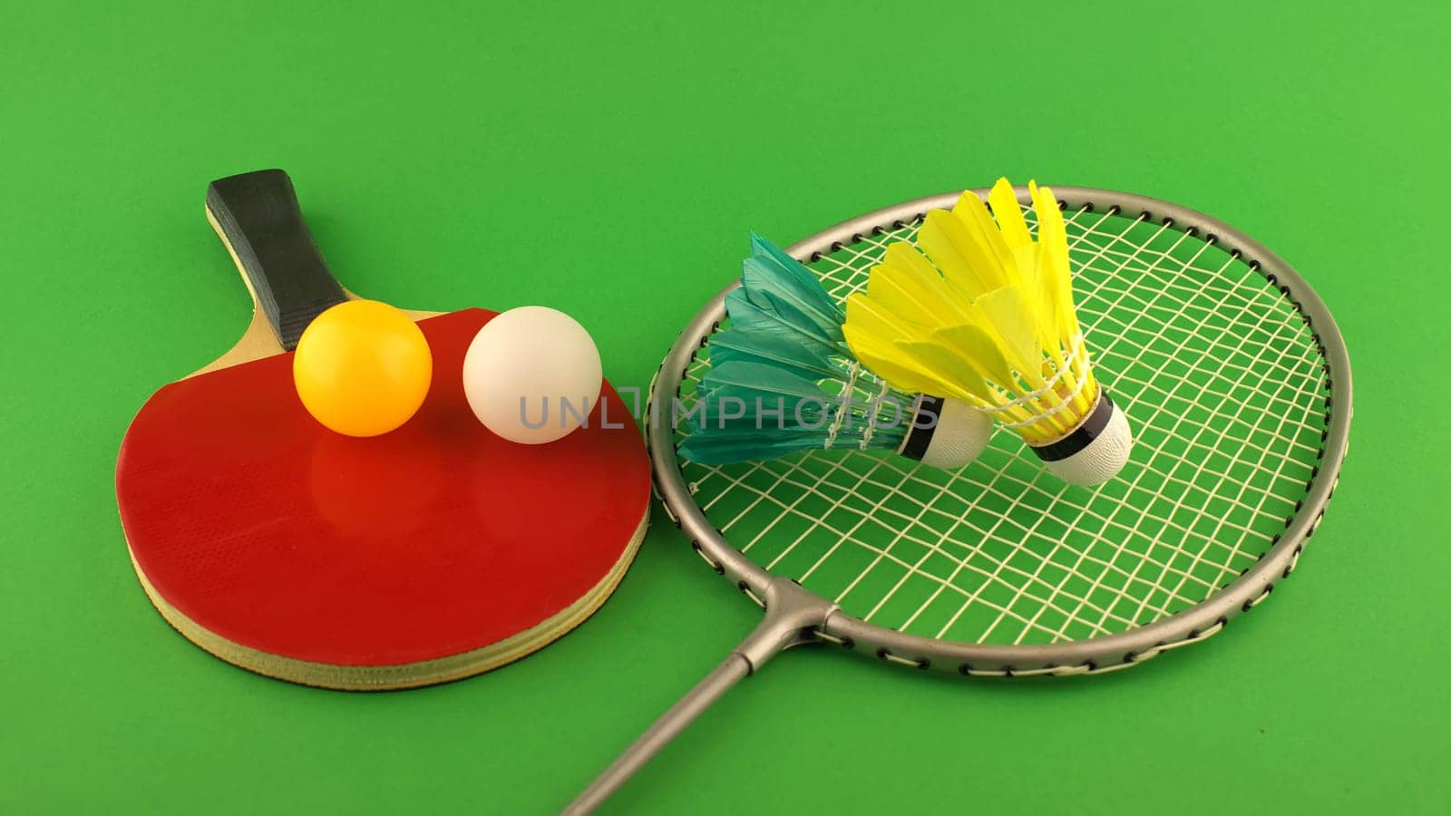 Table tennis racket with a yellow ball and a white ball on it and badminton racket with a feather shuttlecock on clear green background