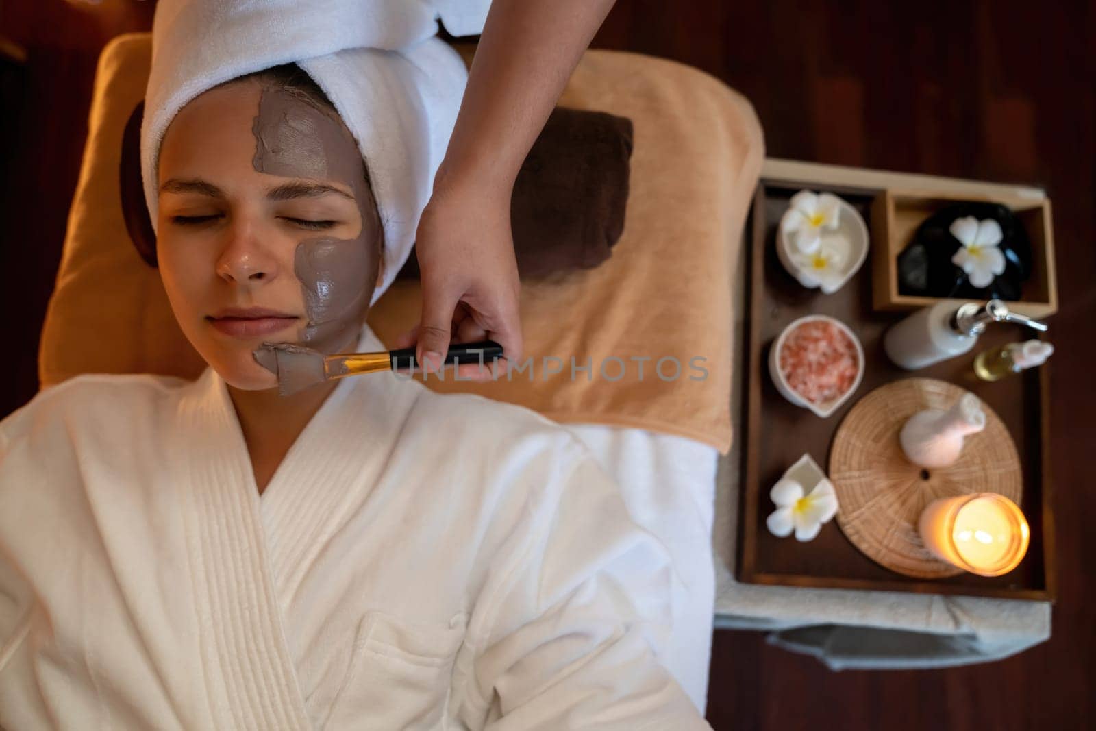 Serene ambiance of spa salon, top view woman customer indulges in rejuvenating with charcoal face cream massage with warm lighting candle. Facial skin treatment and beauty care concept. Quiescent