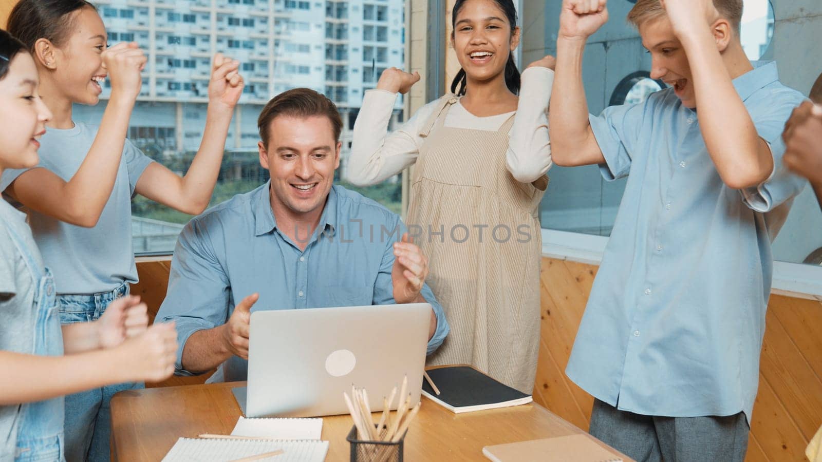 Caucasian teacher and multicultural students clapping hand or putting the hand in the air together to celebrate for successful project with laptop and equipment placed on table in class. Edification.