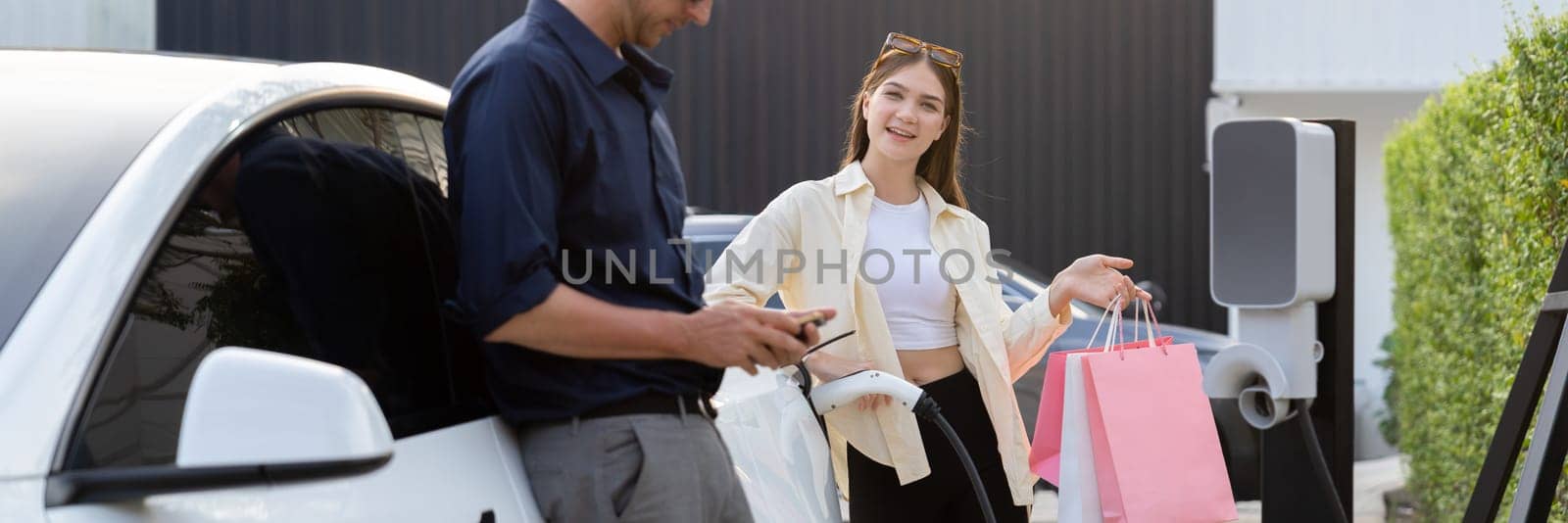 Young couple with shopping bag use smartphone to pay for electricity to recharge EV car battery from charging station at city mall parking lot. Happy couple go shopping by eco car.Panorama Expedient