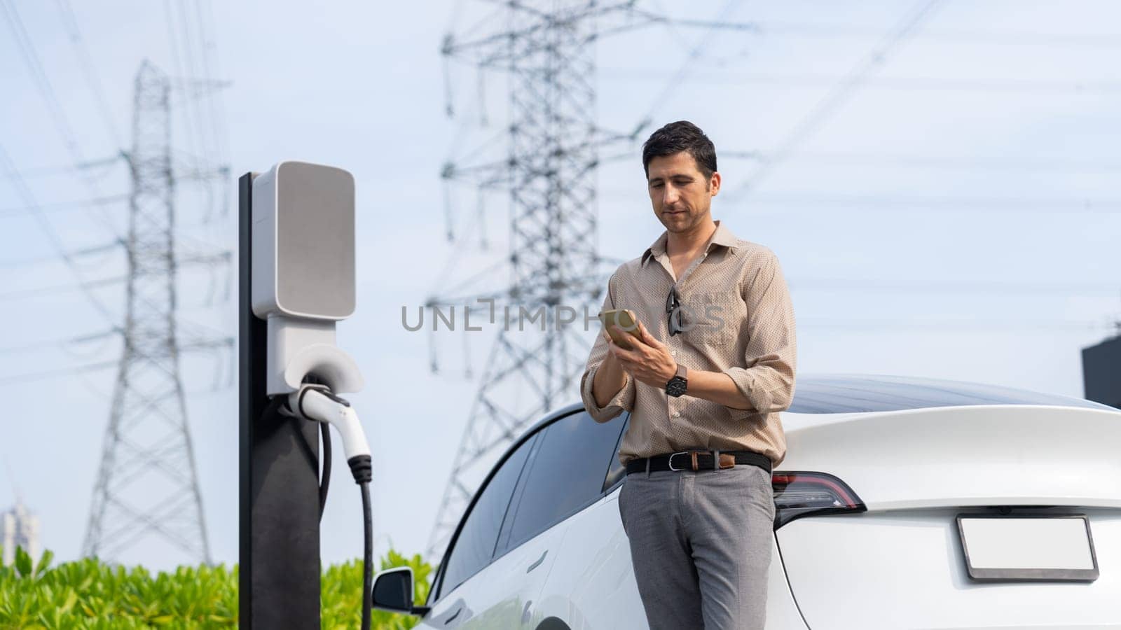 Man pay for electricity with smartphone while recharge EV car battery at charging station connected to electrical lower grid tower for eco friendly car utilization. Panorama Expedient