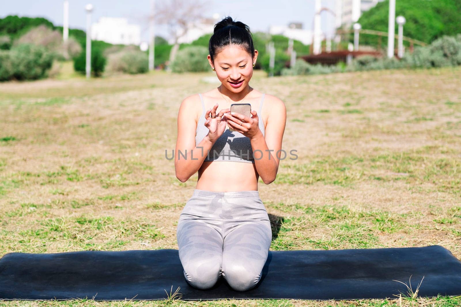 smiling young asian woman in sportswear kneeling on her mat using mobile phone for a yoga session on the park grass, technology and healthy lifestyle concept