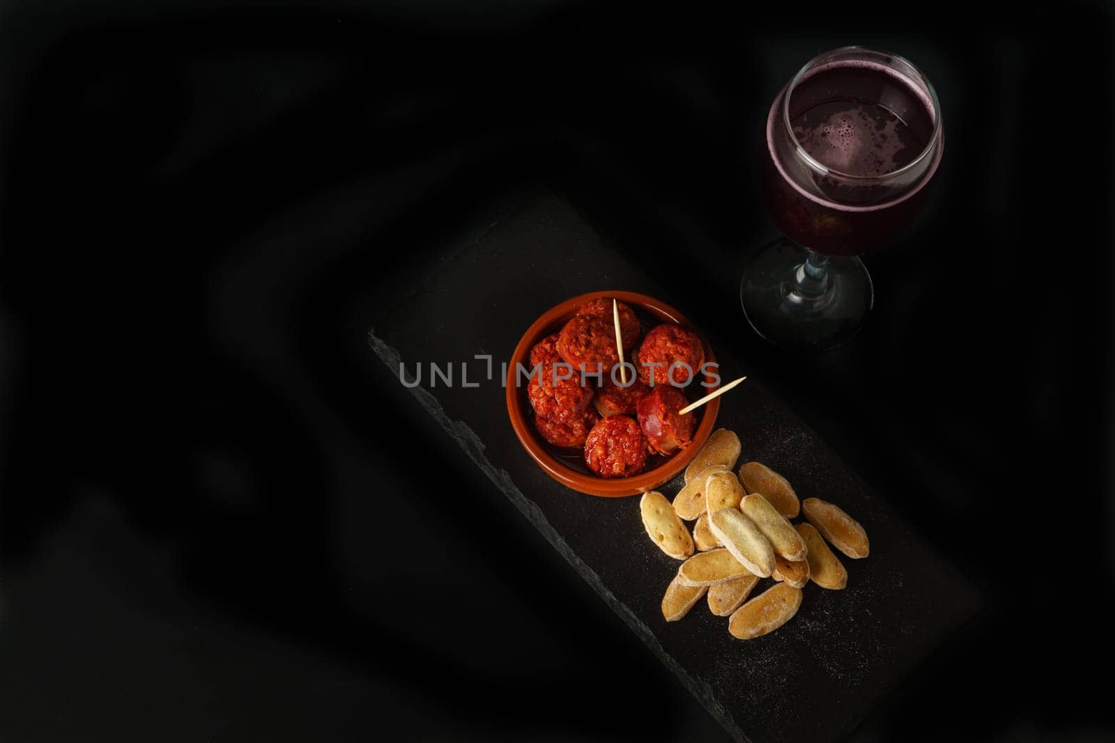 top view of an earthenware casserole with fried sausage, bread and a glass of wine isolated on a black background.