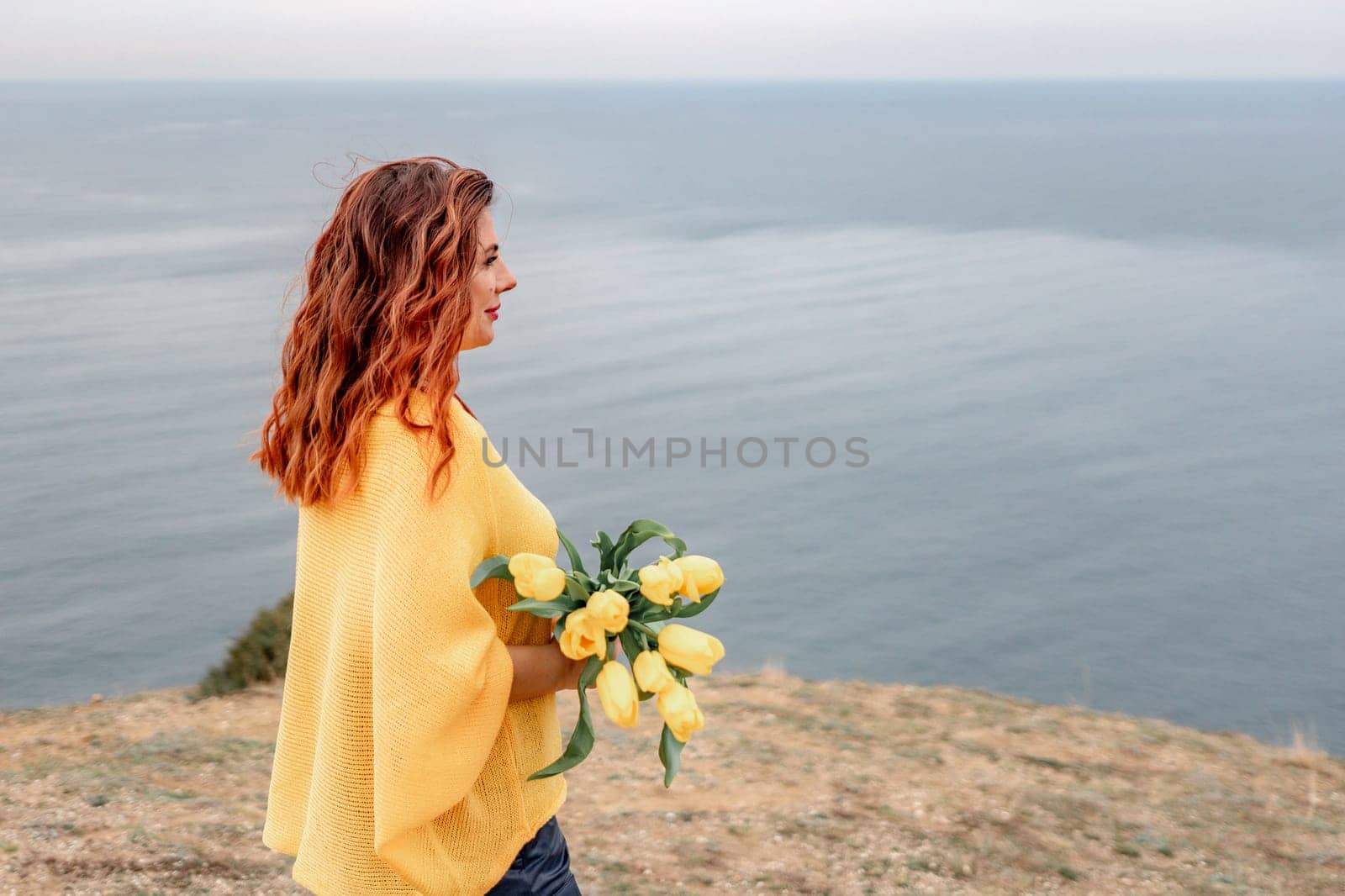 Rear view of a woman with long hair against a background of mountains and sea. Holding a bouquet of yellow tulips in her hands, wearing a yellow sweater.