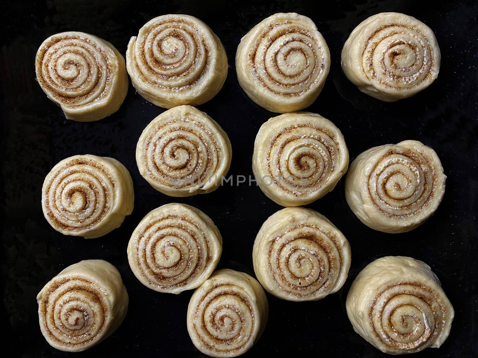 preparing homemade round cinnamon scrolls with sugar crystals in a rectangular black baking sheet, ready for baking.