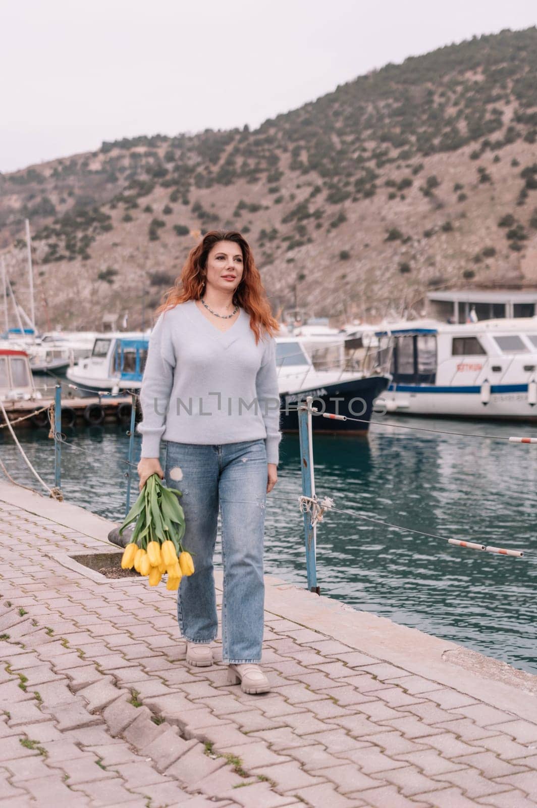 Woman holds yellow tulips in harbor with boats docked in the background., overcast day, yellow sweater, mountains.