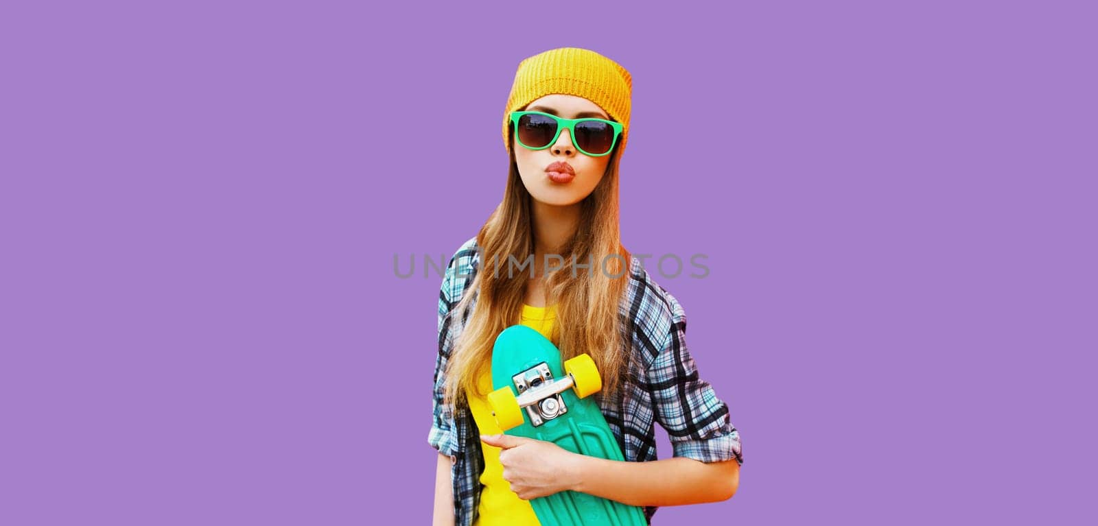 Summer portrait of happy blonde young woman posing with skateboard in colorful clothes on purple studio background