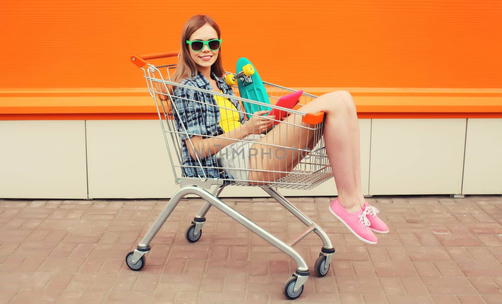 Modern stylish young woman using digital tablet computer sitting in shopping trolley cart on city street