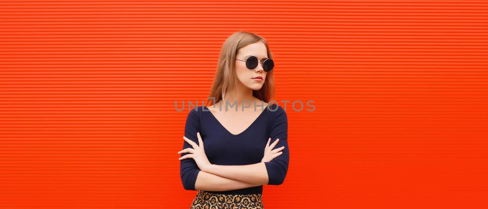 Portrait of beautiful young blonde woman posing in black round sunglasses looking away on red background