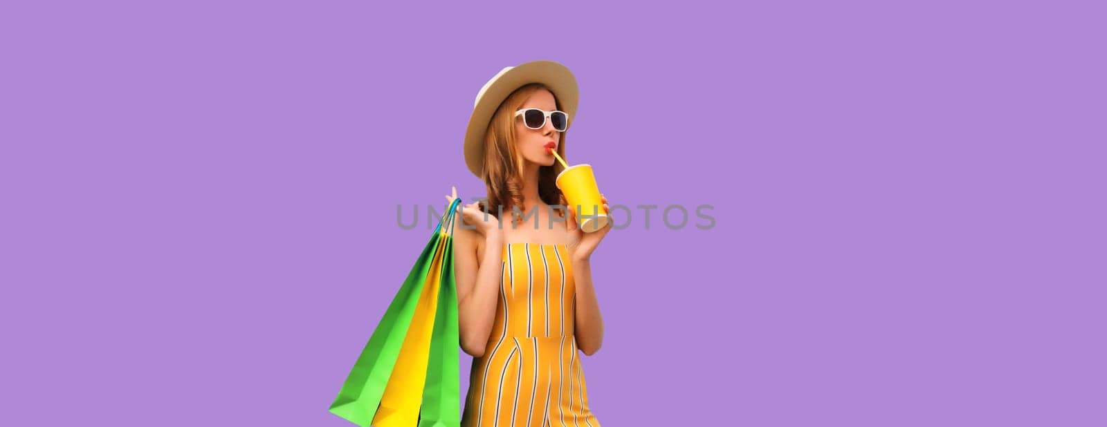 Portrait of stylish beautiful young woman posing with shopping bags drinking coffee or juice in summer hat on purple studio background