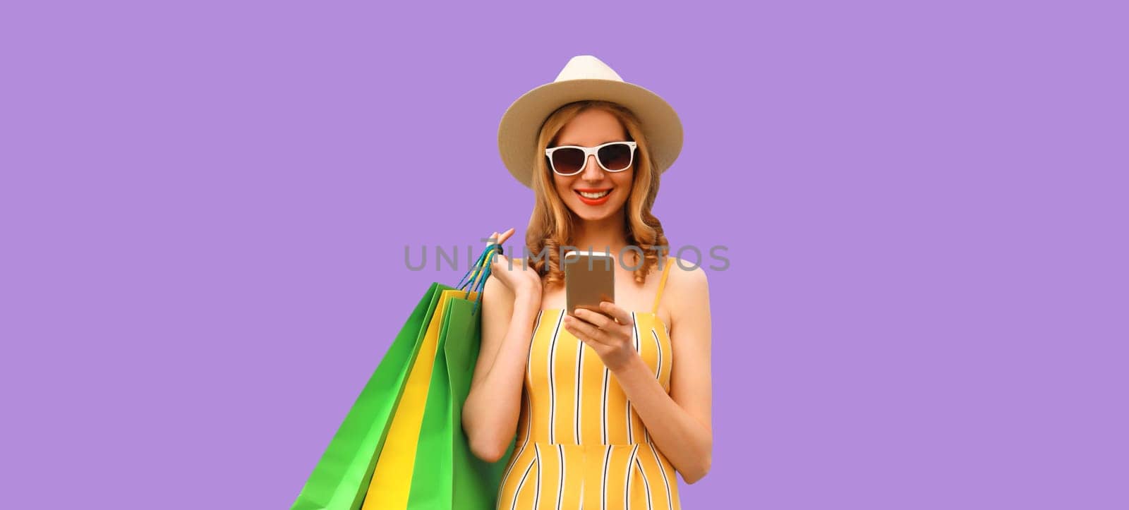 Beautiful happy young woman looking at phone with colorful shopping bag in summer straw hat on purple studio background