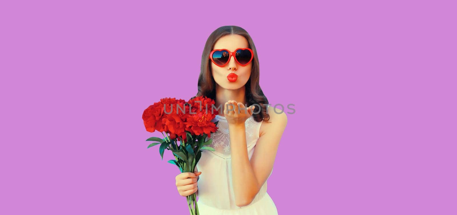 Portrait of beautiful young woman with bouquet of red rose flowers and blowing kiss in heart shaped sunglasses on purple studio background