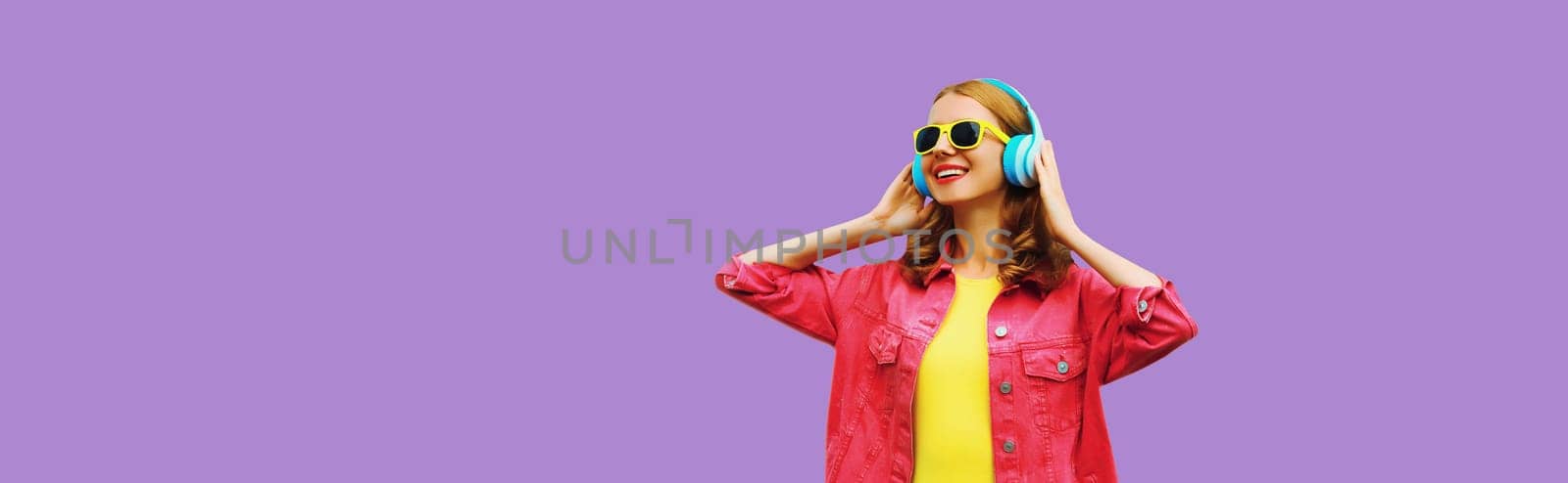 stylish modern happy young woman listening to music with headphones on colorful purple studio background, blank copy space for advertising text