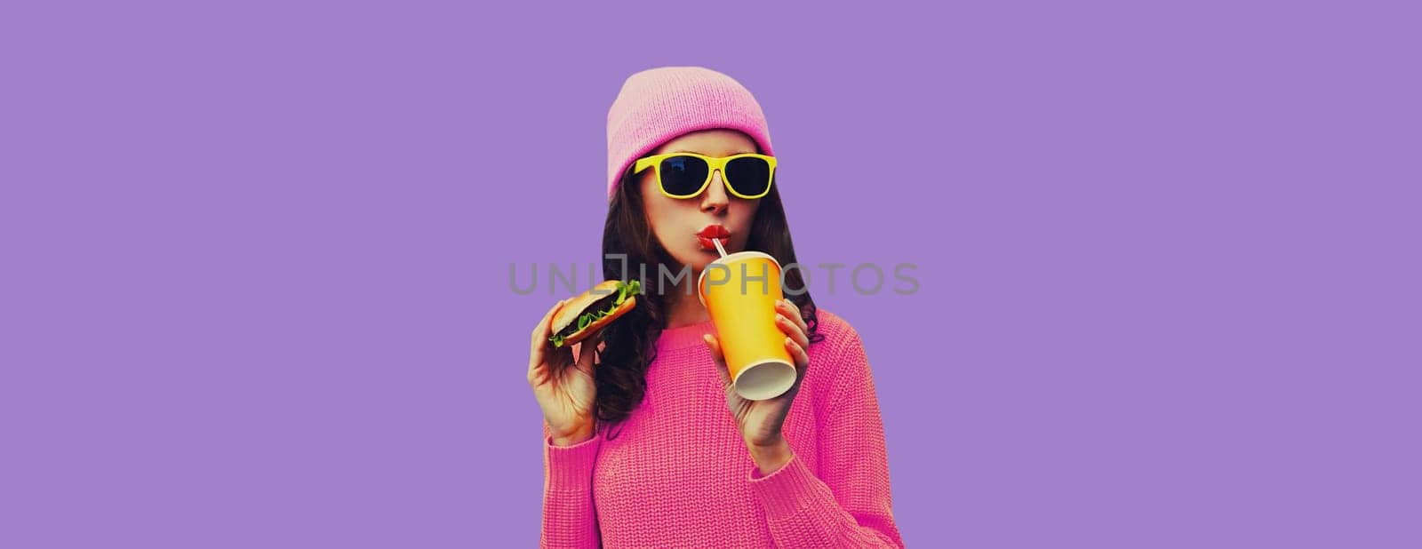 Portrait of stylish modern young woman drinking juice eating burger fast food on colorful purple studio background