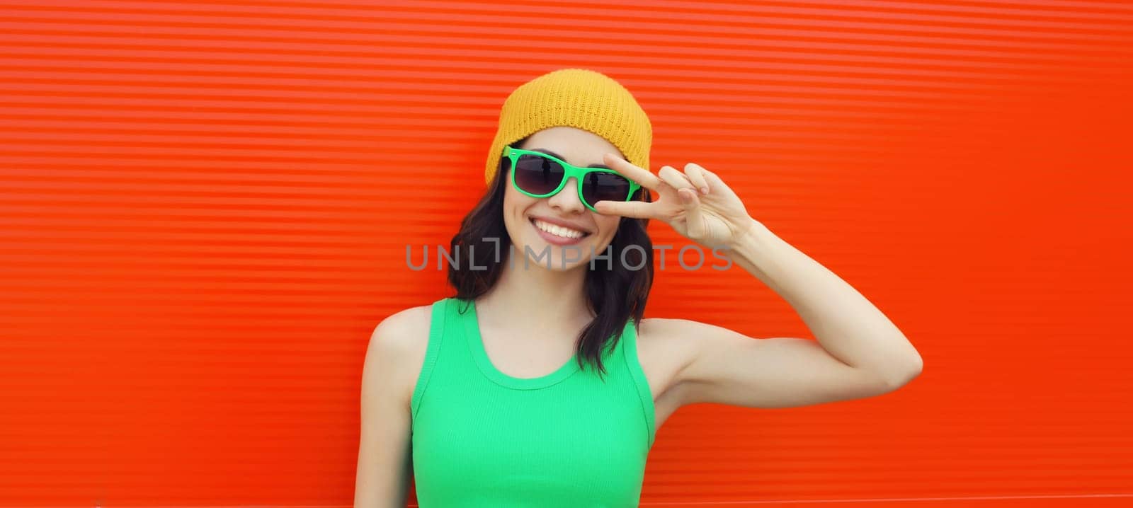 Summer portrait of happy smiling brunette young woman posing in yellow hat, green sunglasses on red background