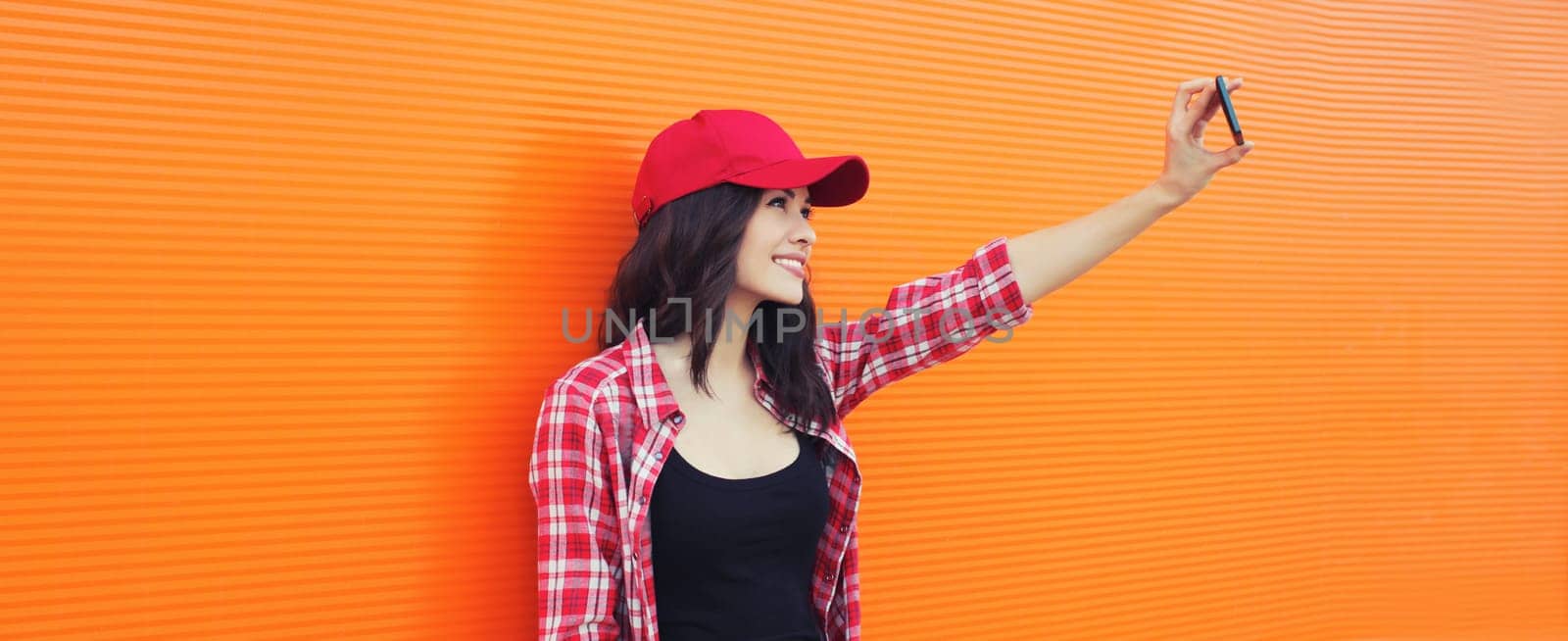 Summer portrait of happy smiling young woman taking selfie with mobile phone in red baseball cap on colorful orange background on city street