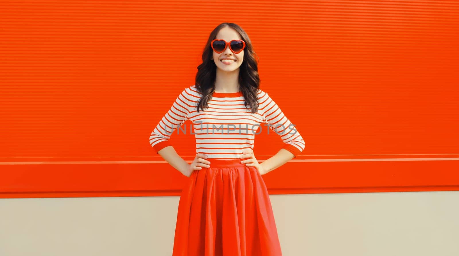 Beautiful happy smiling young brunette woman model posing in red heart shaped sunglasses, skirt on city street