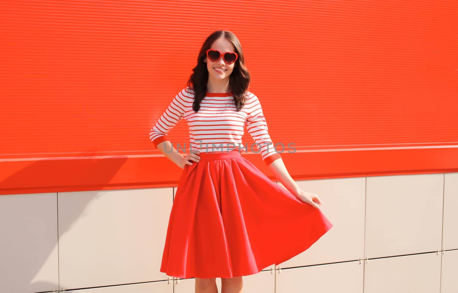 Beautiful happy smiling young brunette woman model posing in red heart shaped sunglasses, skirt on city street
