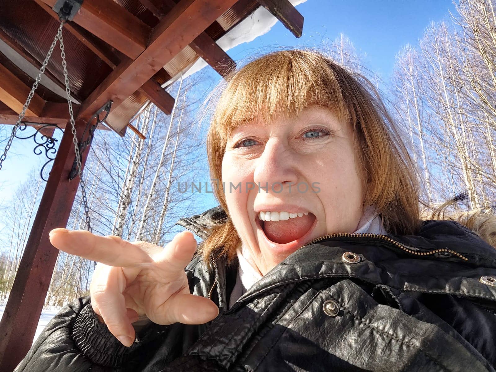A cheerful middle aged woman in a winter coat taking selfie on the swing on nature outdoors in sunny day with blue sky