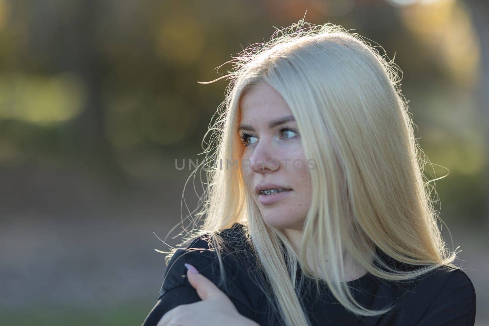 A young woman with blonde hair and braces is smiling while wearing a black shirt in a park setting.