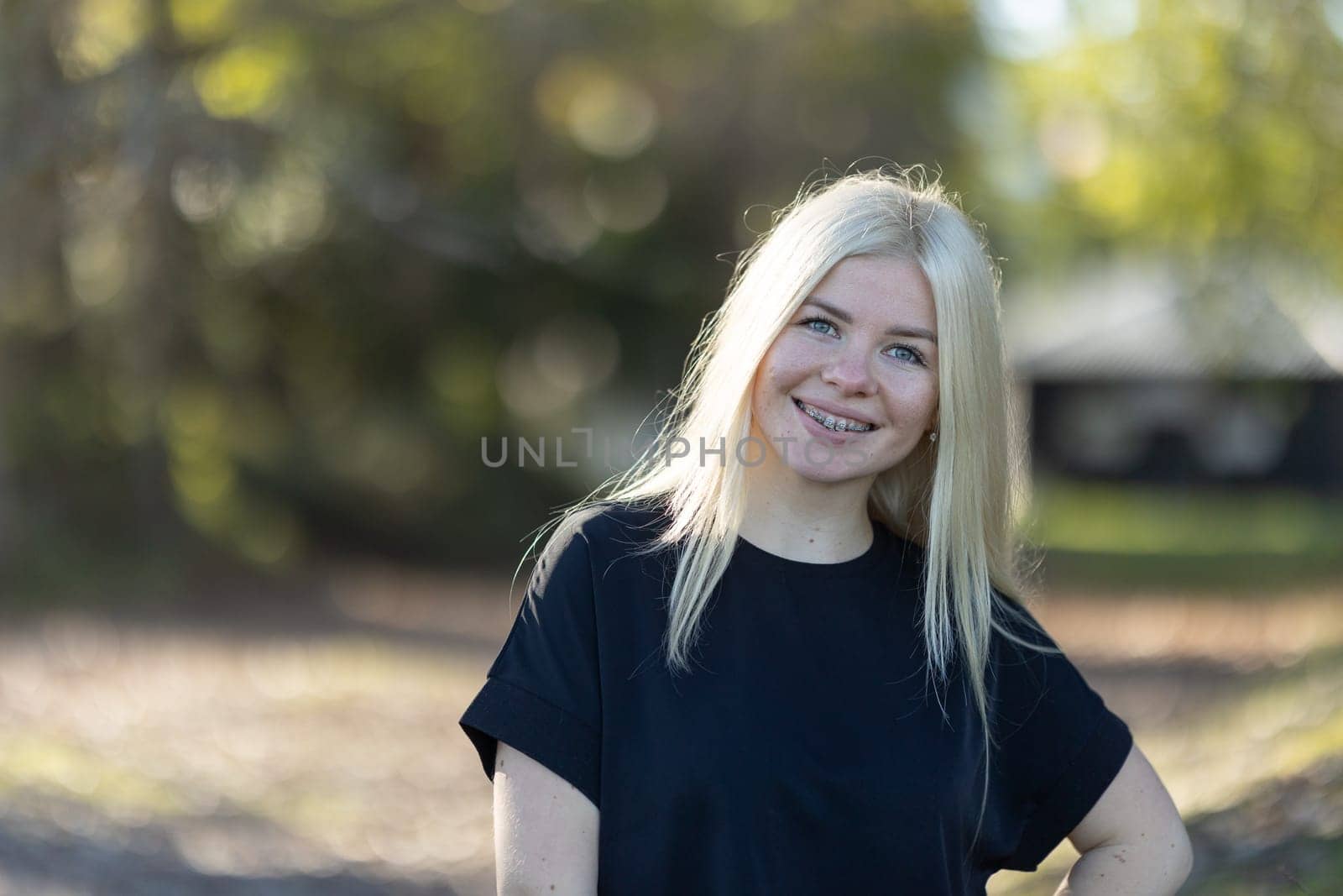 A young woman with blonde hair and braces is smiling while standing in a park surrounded by greenery and trees on a sunny day.