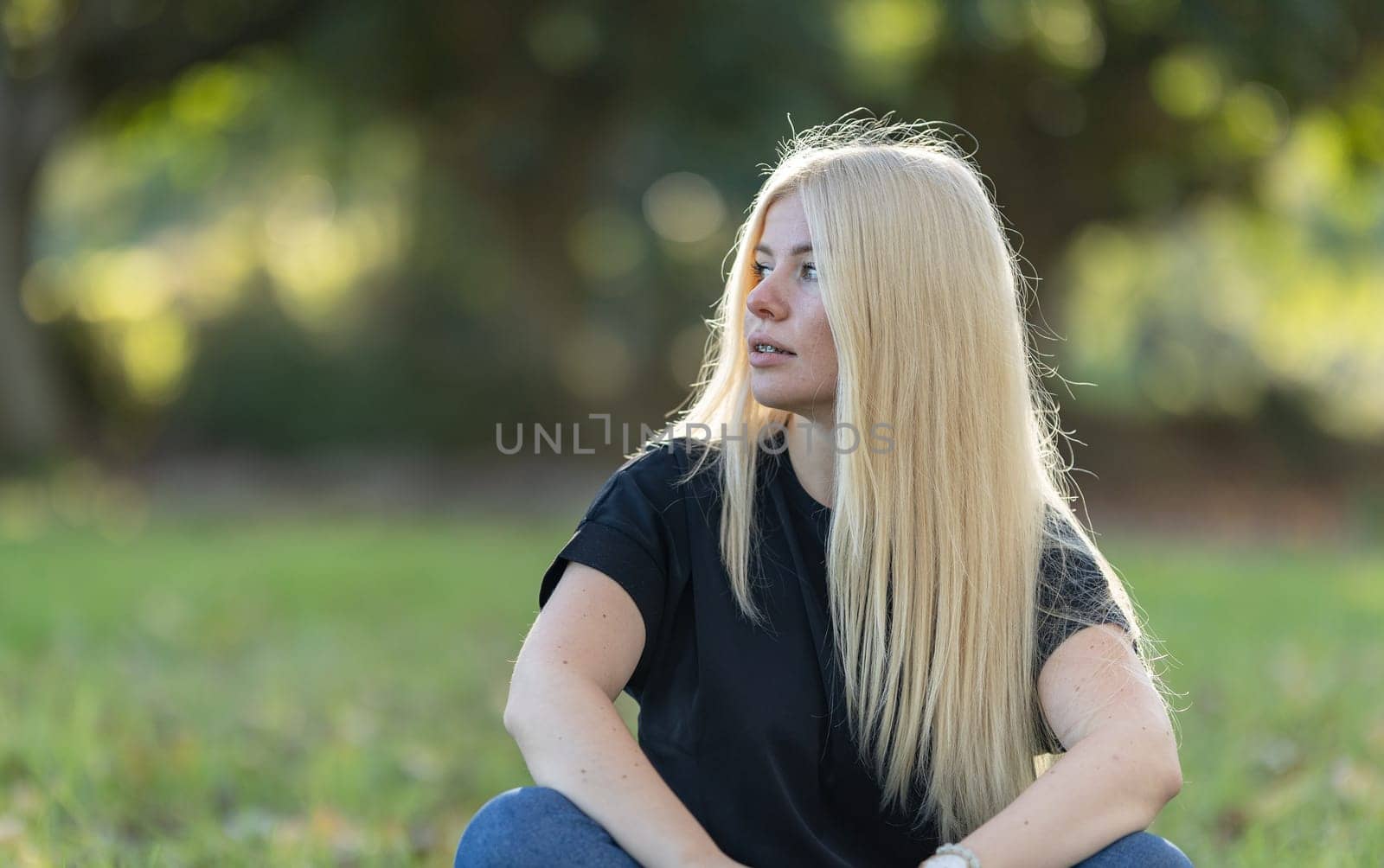 A young woman with long blonde hair and braces is sitting on the grass in a park, smiling.