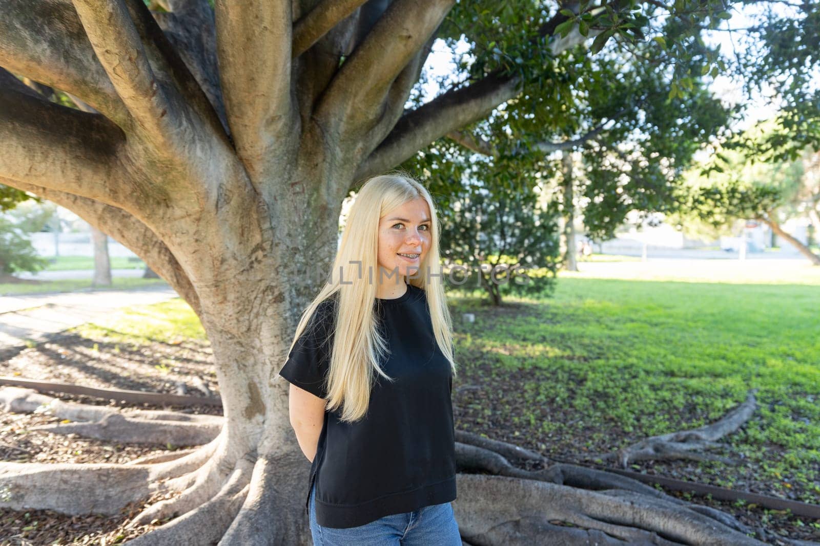 A young woman with braces is standing in front of a tree in a park, smiling.