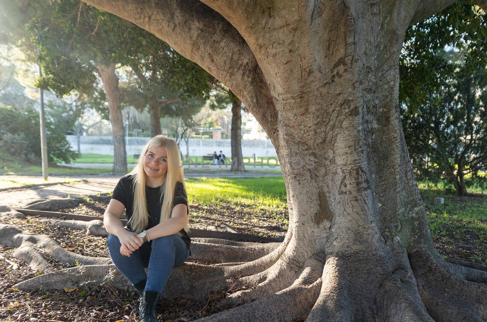 A young woman with braces is sitting under a large tree in a park, smiling and enjoying the outdoors on a sunny day.