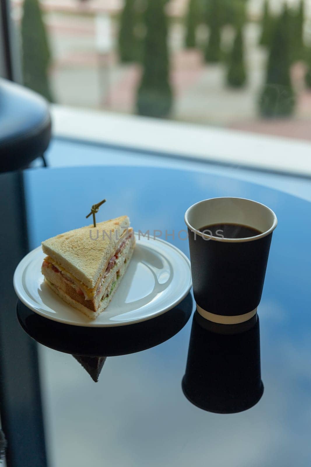 Paper cup of coffee and sandwich in the garden on a glass table with a reflection of the sky. Vertical image.