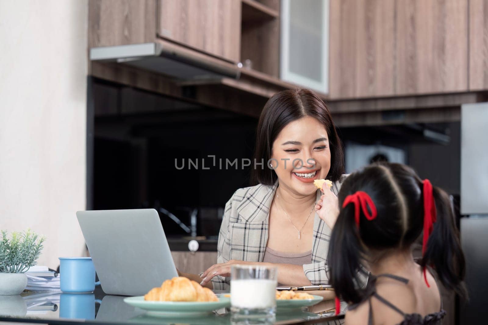 Working mom work from home office. Happy mother and daughter feed snacks to each other. businesswoman and cute child using laptop work freelancer workplace in home, Lifestyle family moment.