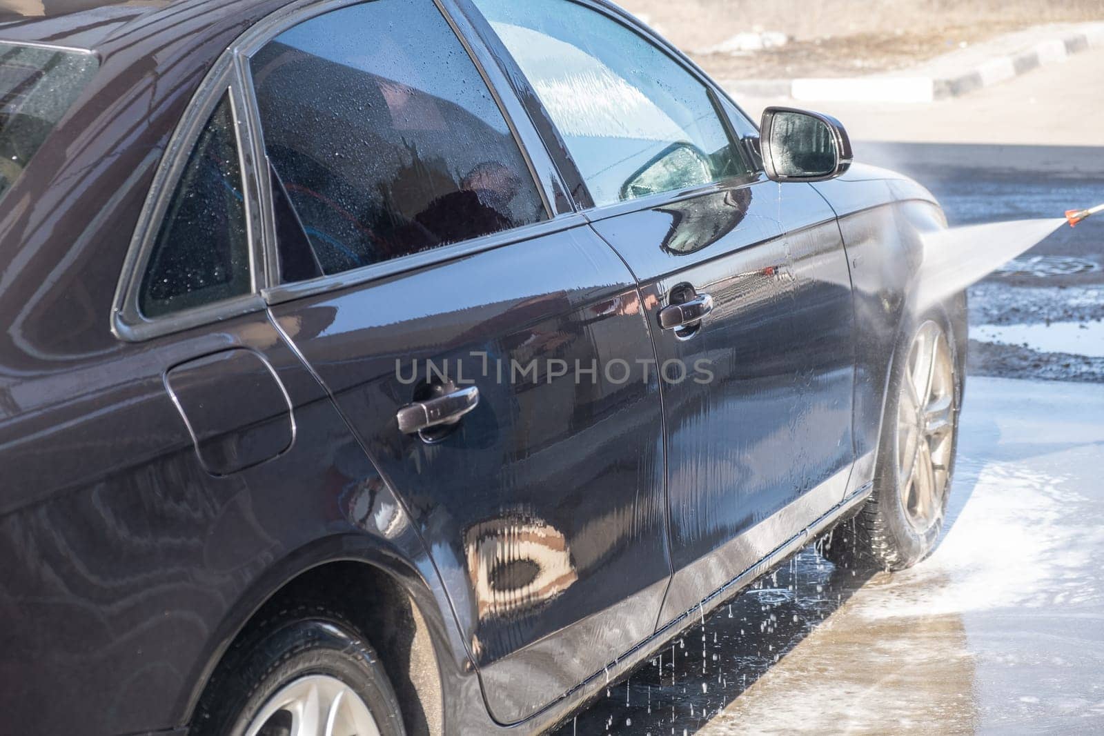 A man washes off the foam while washing the car. A vehicle is engulfed in soapy foam during a car wash, covering the tires, wheels, hood, and entire exterior of the car. Self-service car wash