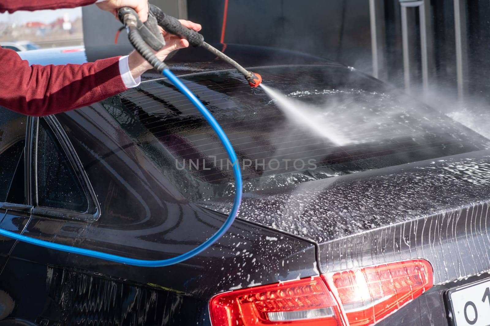 A man washes off the foam while washing the car. A vehicle is engulfed in soapy foam during a car wash, covering the tires, wheels, hood, and entire exterior of the car. Self-service car wash