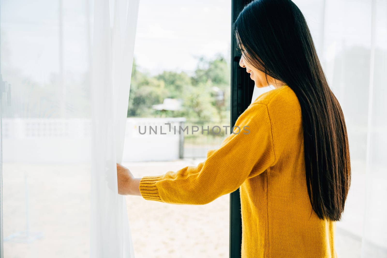 A young woman opens curtains in the early morning smiles at the view feeling relaxed by Sorapop