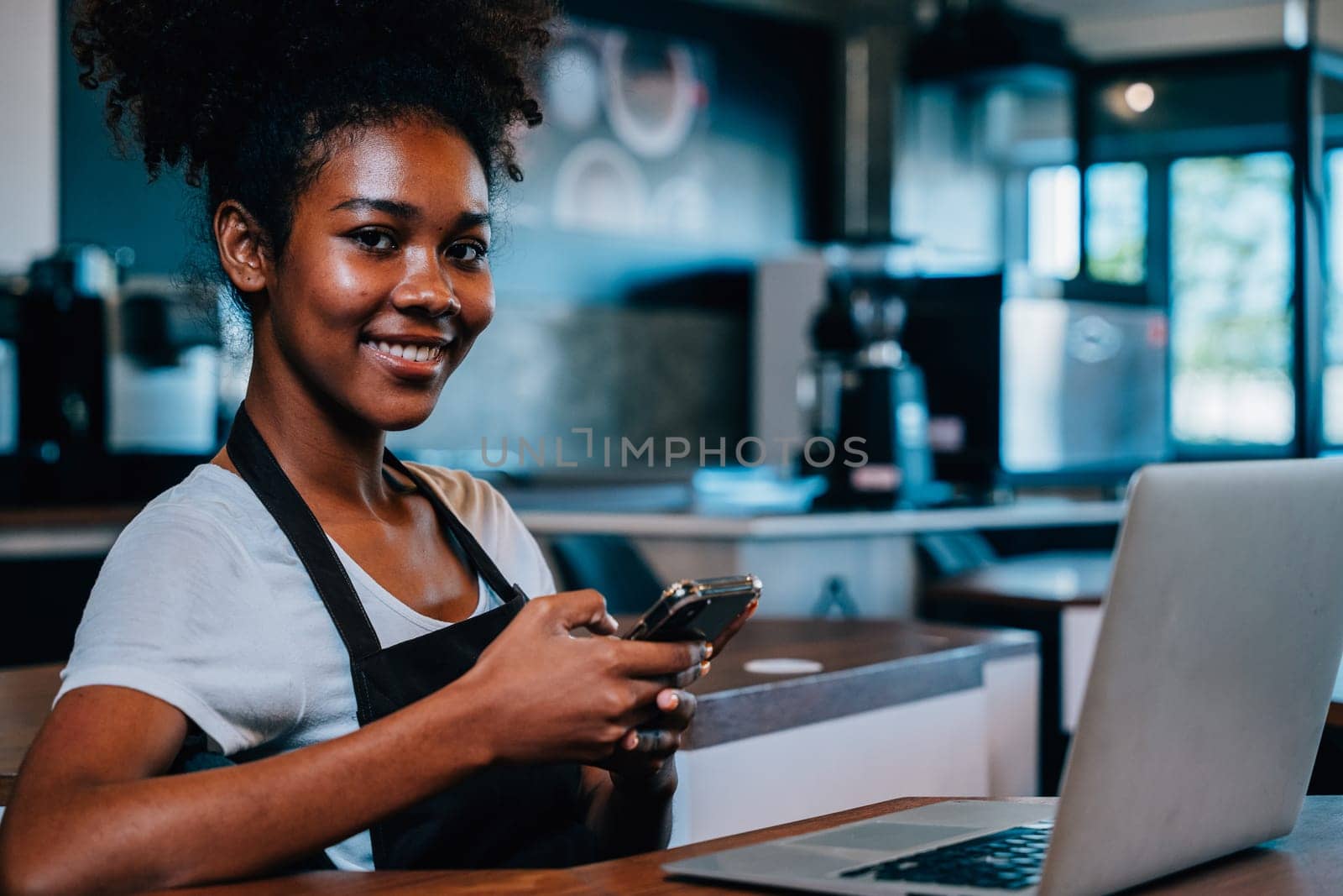 Coffee shop owner a focused black woman in apron using smartphone in her cafe. Multi tasking barista takes orders communicates with customers manages her successful business. by Sorapop