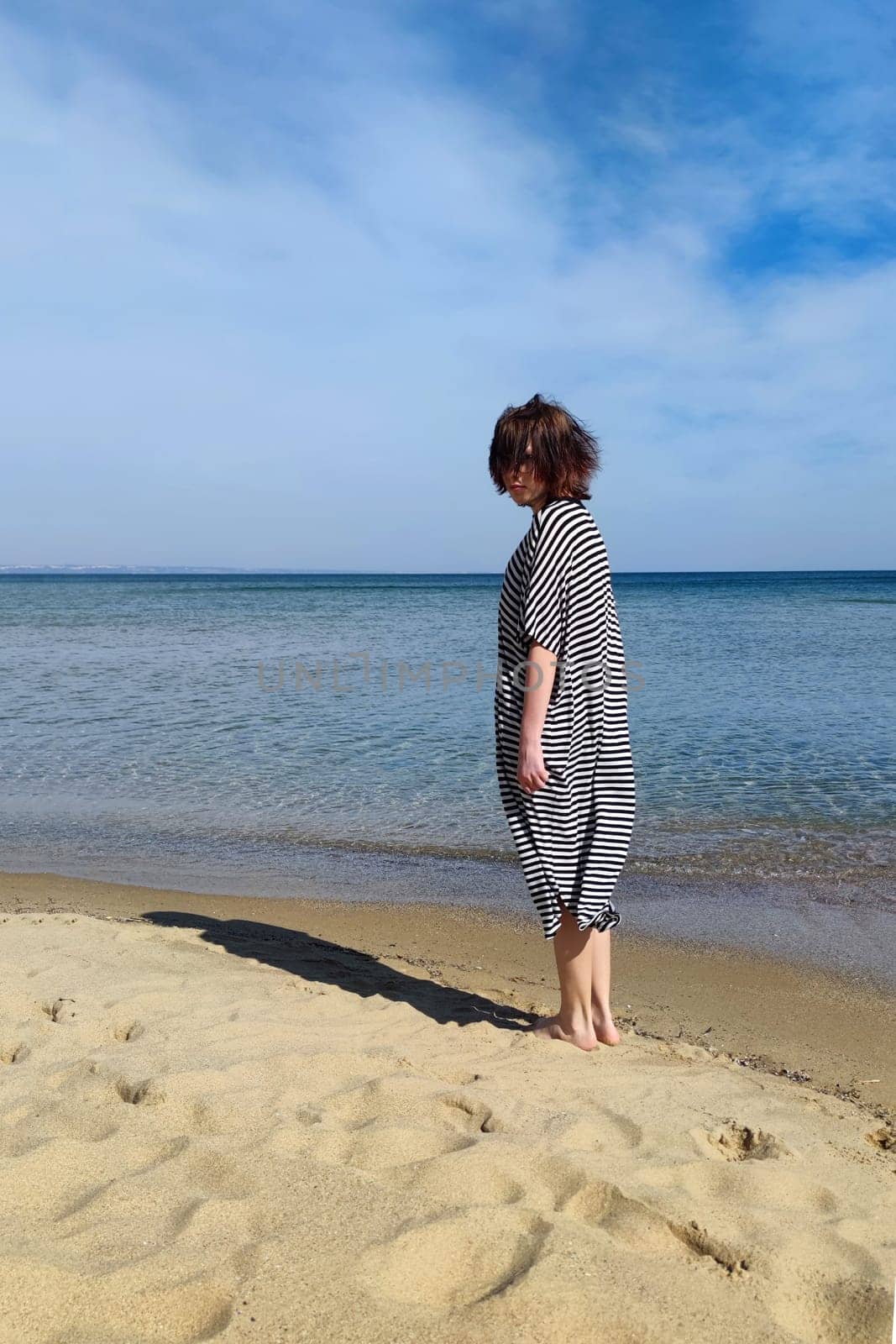 teenage girl with brown hair in a striped dress barefoot on the seashore on a sunny day