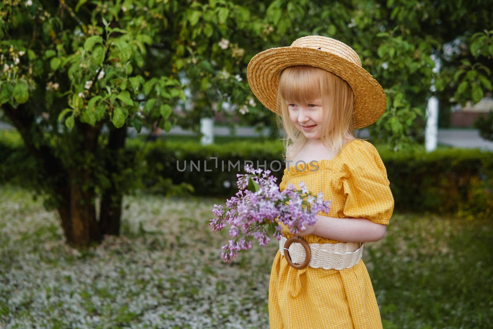 A little girl in a yellow dress and straw hat wearing a bouquet of lilacs. A walk in a spring park, blossoming lilacs