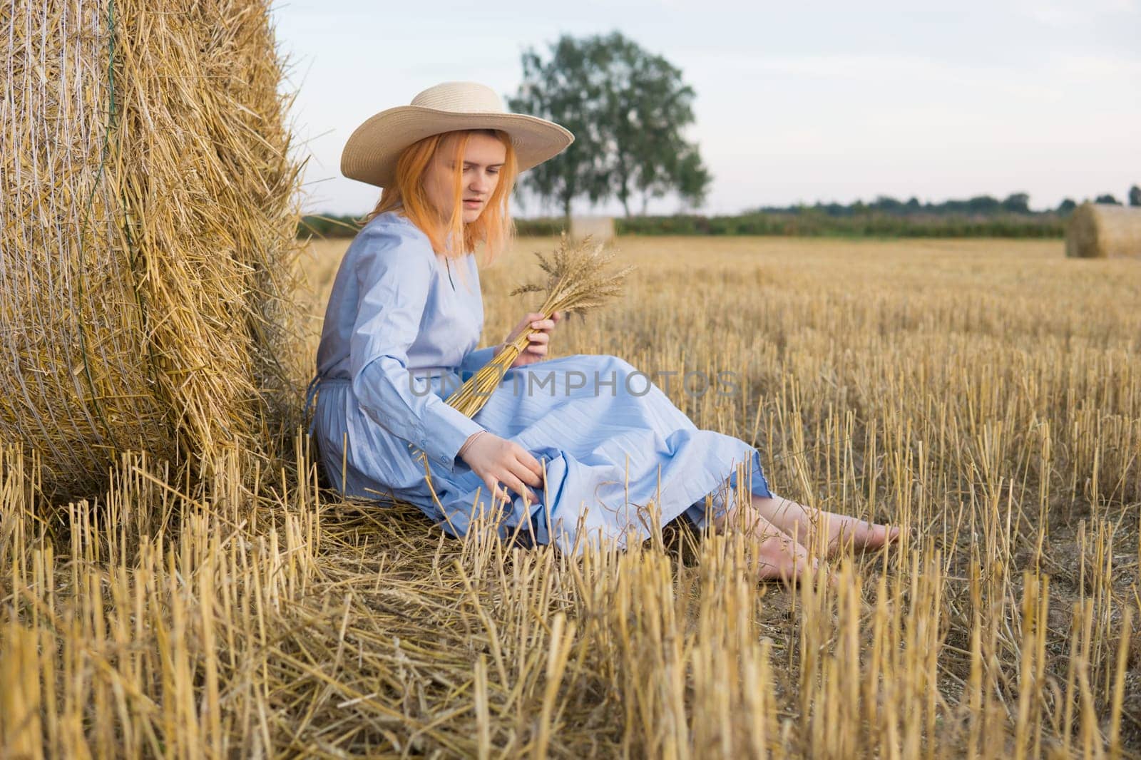 A red-haired woman in a hat and a blue dress walks in a field with haystacks