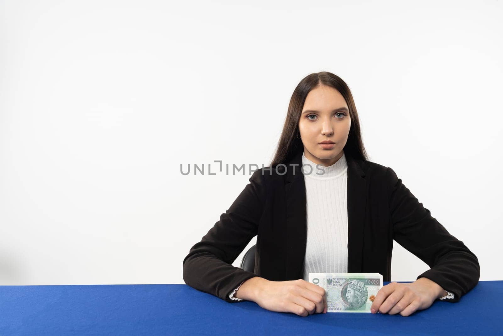 A young brunette holds in front of her a stack of banknotes with a face value of one hundred Polish zlotys. A girl is sitting in a chair at the table with a blue tablecloth. by fotodrobik