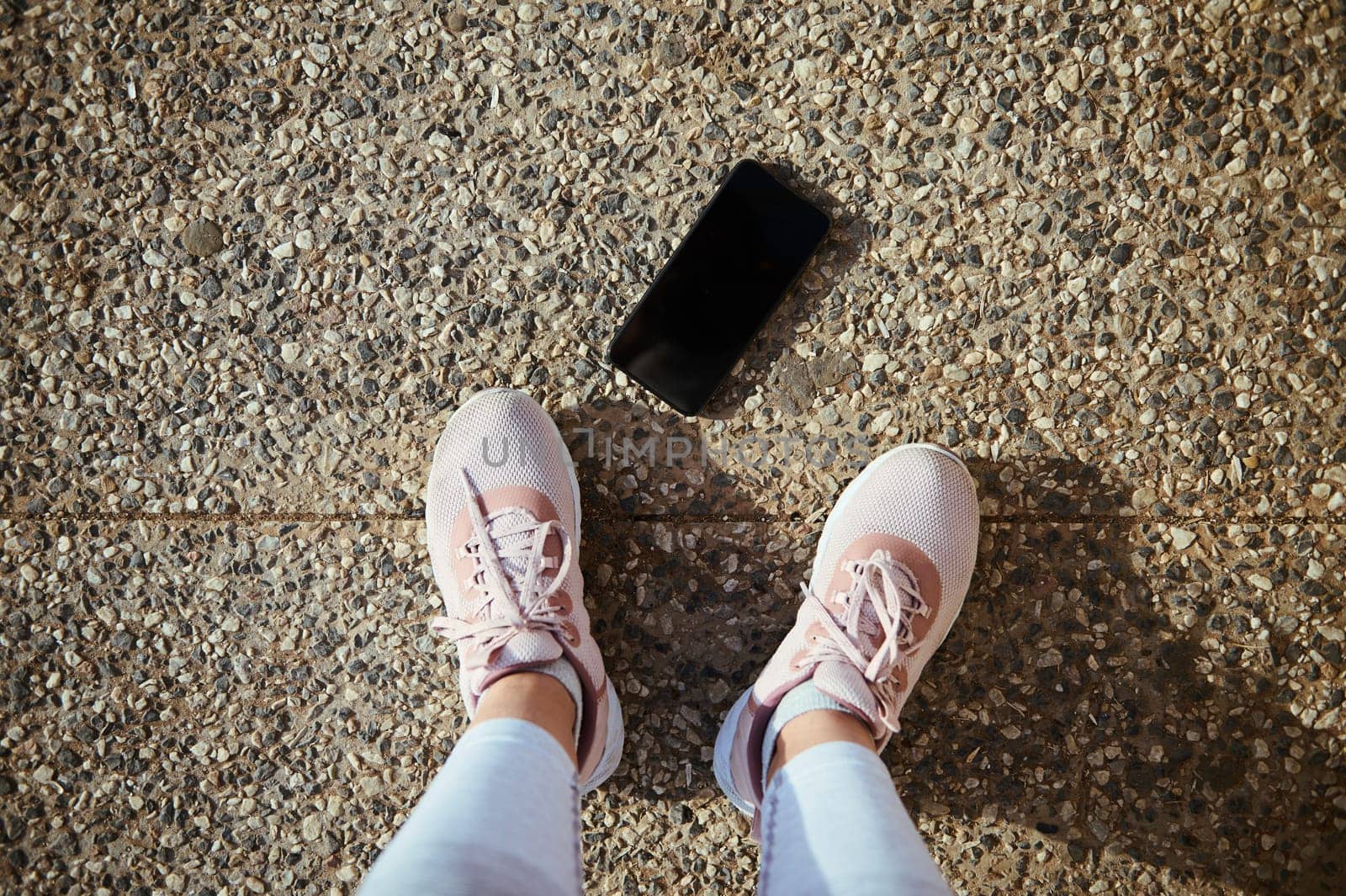 Sportswoman POV. Legs and feet of female athlete in pink sneakers and smartphone with mockup black screen on the asphalt by artgf