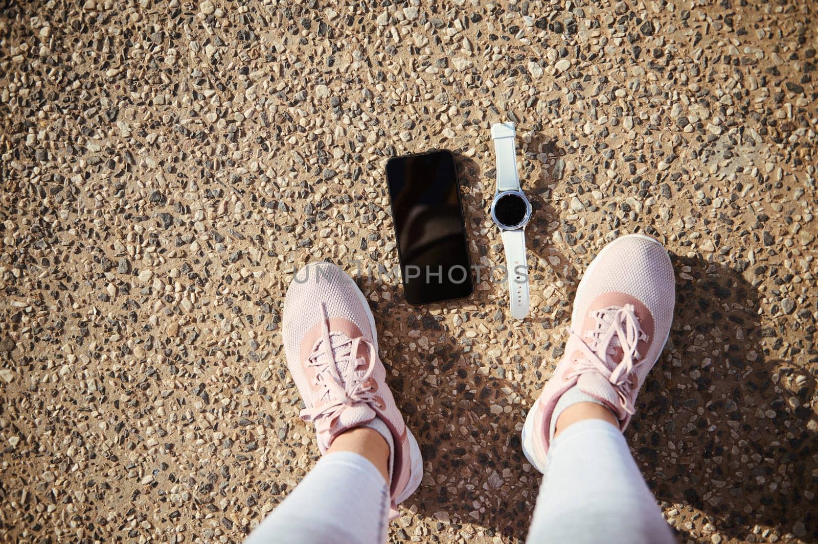 Top view feet of a sportswoman in pink running sports shoes, a smart wrist watch and smartphone with black blank mockup digital screen on the asphalt. Copy ad space for sports and fitness mobile app