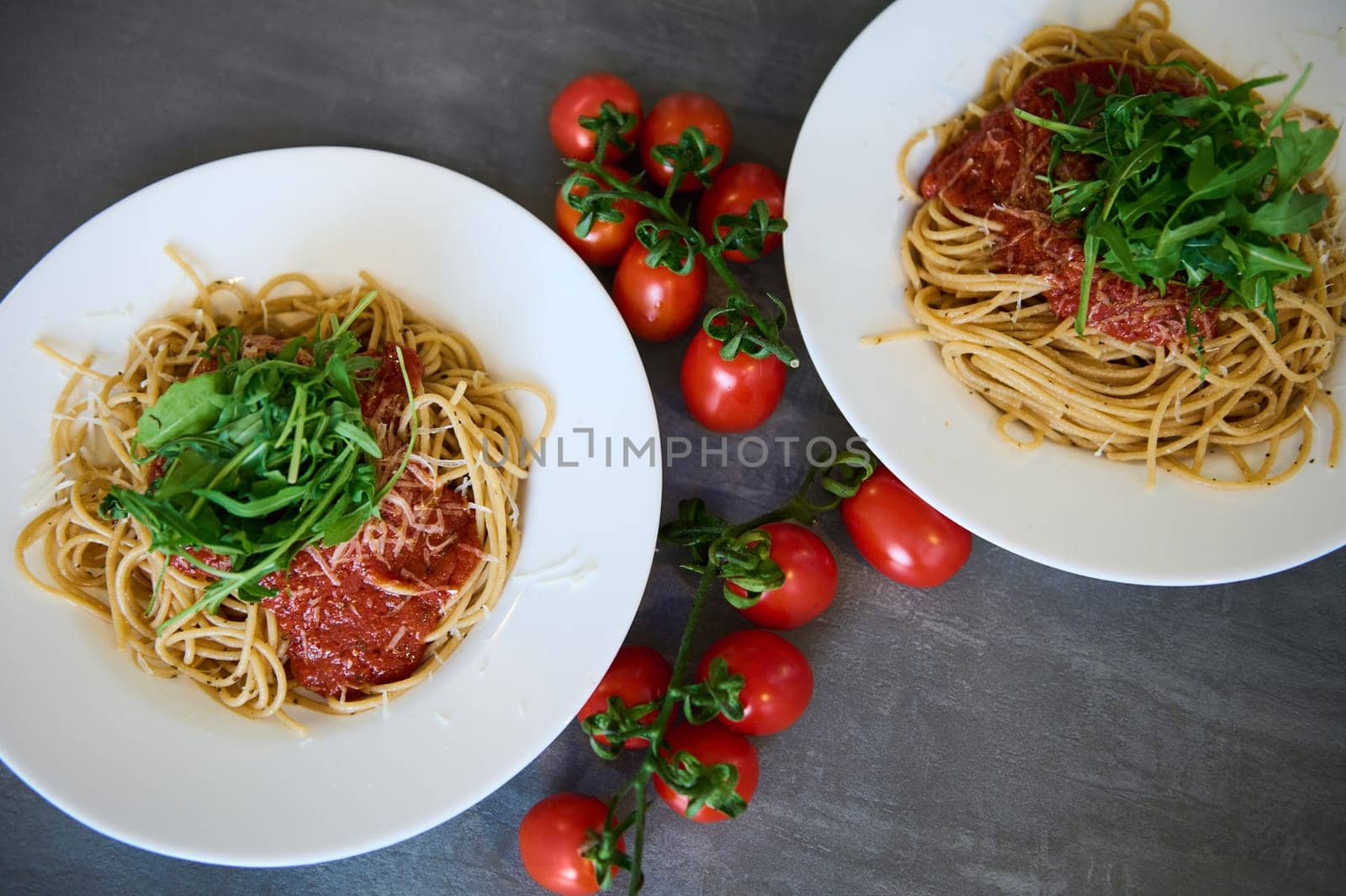 Top view white plates of Italian pasta, sauce and green arugula leaves next to sprig of cherry tomatoes on a gray table by artgf