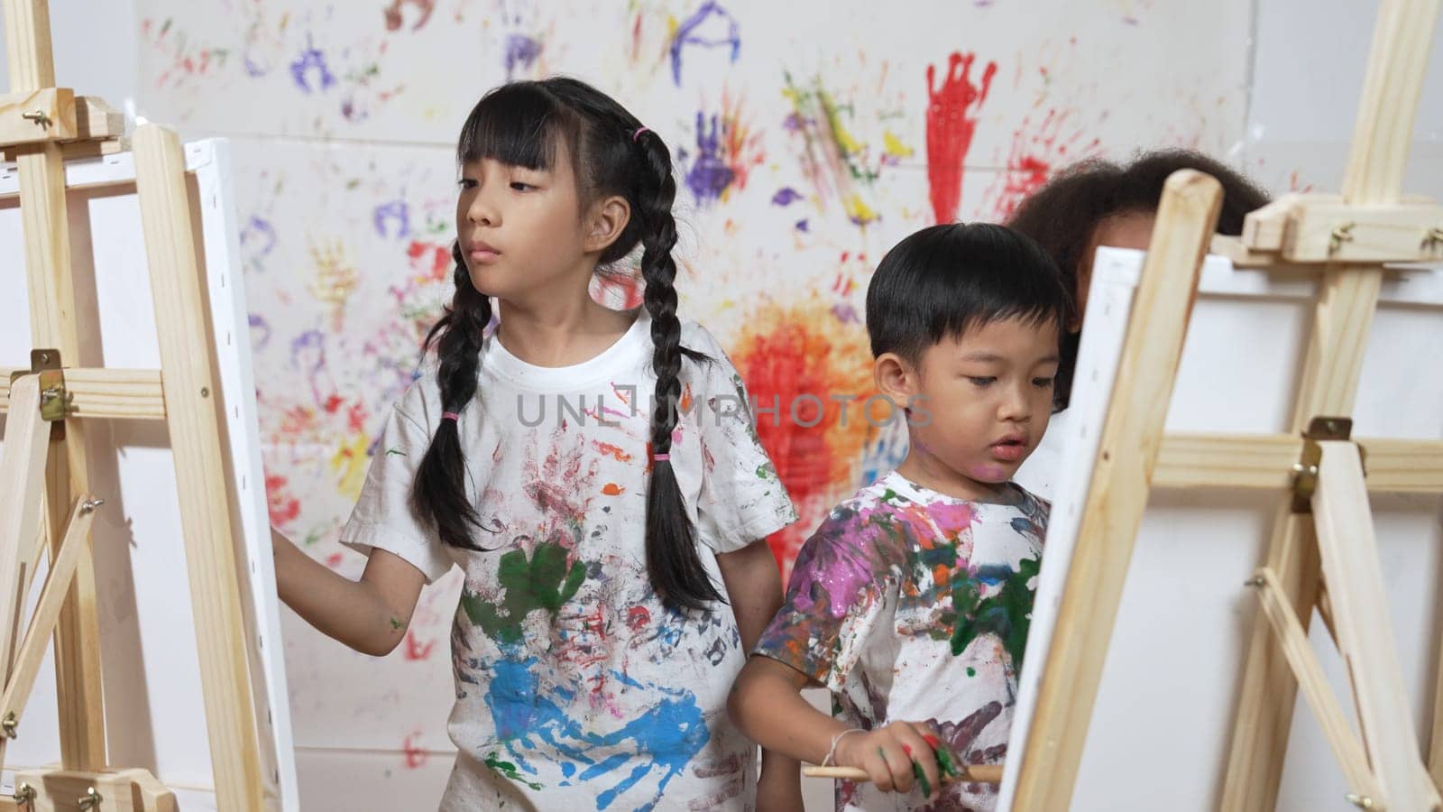 Playful student painted or draw canvas at stained wall with diverse friends in art lesson. Group of happy multicultural children working or create artwork at messy room. Creative activity. Erudition.