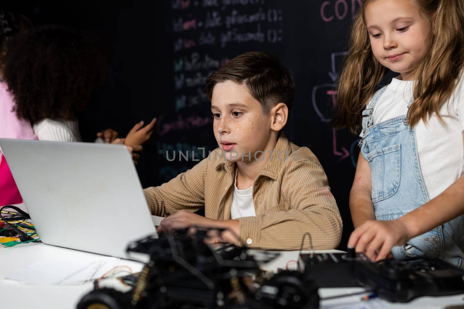 In classroom student in STEM class learning about coding robotics car. Brown shirt Schoolboy and white bib schoolgirl watching laptop and discussing. Other schoolgirls playing around funny. Erudition.